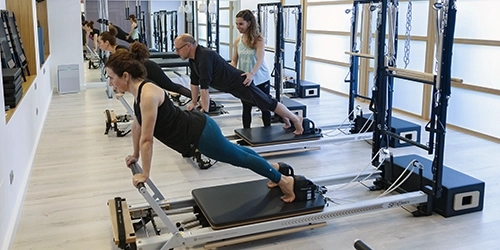 Cuatro alumnos haciendo ejercicios de pilates en maquinas reformer mientras una monitora corrige las posturas en una sala de Esencia Pilates