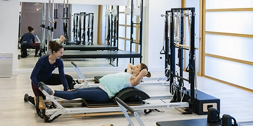 Mujer embarazada realizando ejercicios de pilates en una máquina reformer supervisada por una monitora en una sala de Esencia Pilates
